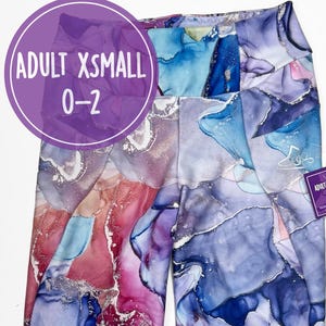 Könnte beinhalten: Hoch taillierte Leggings mit einem abstrakten Alkoholtintenmuster in Blau-, Lila-, Rosa- und Weißtönen mit silbernen Akzenten. Die Leggings sind mit Adult Xsmall 0-2 gekennzeichnet und enthalten ein passendes Stoff-Haargummi mit einem kleinen Etikett.