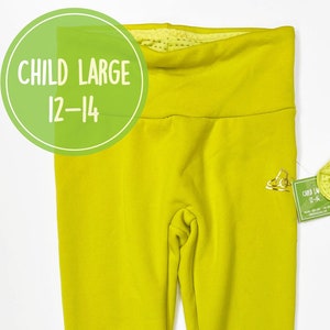 Könnte beinhalten: Eine hellgelbe Leggings mit einem kleinen goldenen Logo am rechten Bein. Die Leggings ist gefaltet und flach auf einem weißen Hintergrund gelegt. Der Text "CHILD LARGE 12-14" ist in einem grünen Kreis sichtbar.