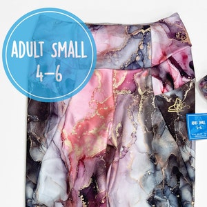 Könnte beinhalten: Eine Leggings in Marmoroptik in den Farben Rosa, Grau und Gold mit einer kleinen Tasche am rechten Bein. Die Leggings ist in der Mitte gefaltet und hat ein blaues Etikett mit der Aufschrift "Adult Small 4-6".