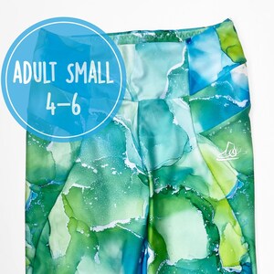 May include: Adult small size leggings with a green, blue, and white abstract watercolor print. The leggings have a small logo on the right leg. The text "ADULT SMALL 4-6" is on a blue circle.