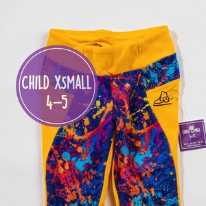 Könnte beinhalten: Gelbe Leggings mit einem bunten Farbklecksmuster. Die Leggings haben einen gelben Bund und ein Seitenpanel mit einem Logo. Das Größenetikett lautet "Child XS-Small 4-5".