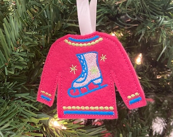 May include: A pink felt Christmas ornament shaped like a sweater with an embroidered ice skate design. The ornament has a white ribbon for hanging.