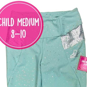 Könnte beinhalten: Hell-türkisfarbene Leggings mit silbernem Sprenkelmuster und passendem Haargummi. Die Leggings haben einen hohen Bund und silberne Bündchen an den Knöcheln. Ein rosa Etikett am Bund trägt die Aufschrift CHILD MEDIUM 8-10, passend zum Kreis.