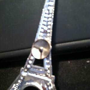 May include: A silver Eiffel Tower charm with a small, round, silver pin on the back.
