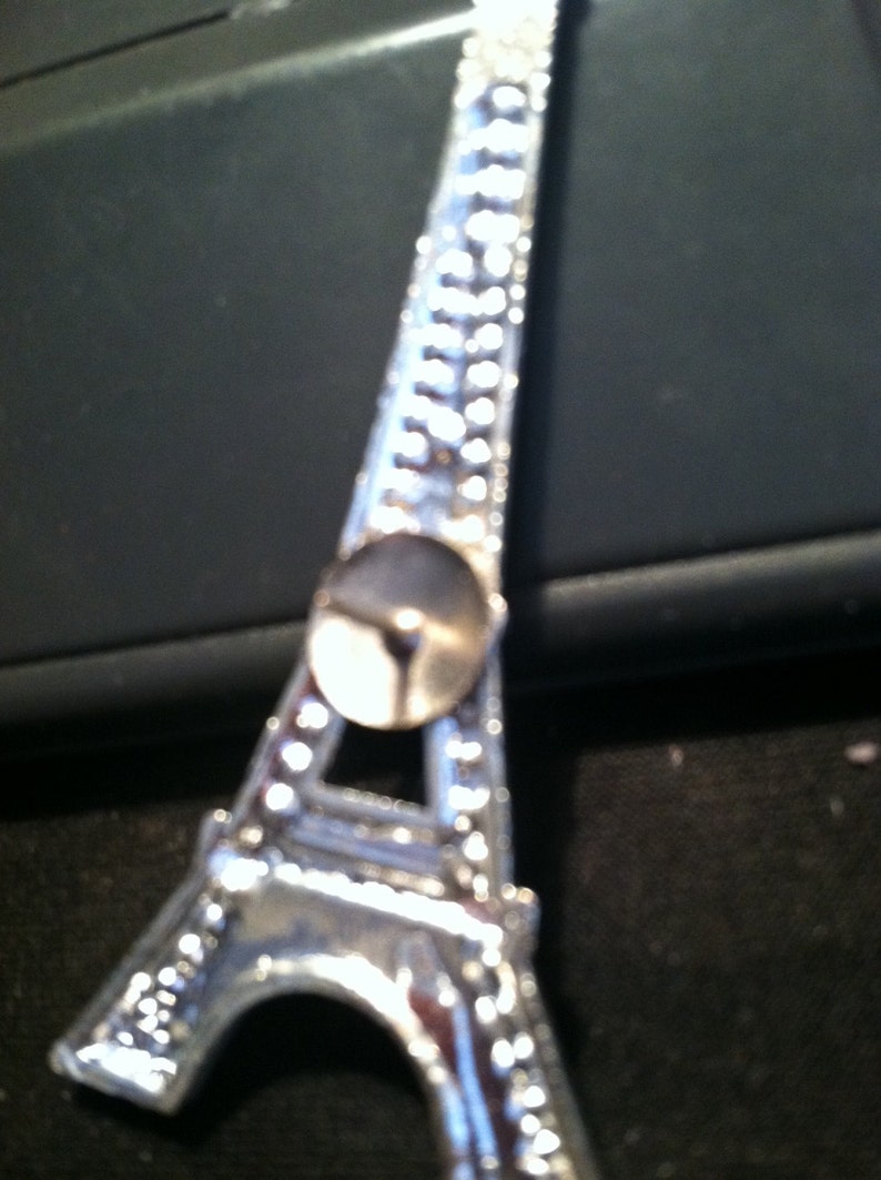 May include: A silver Eiffel Tower charm with a small, round, silver pin on the back.