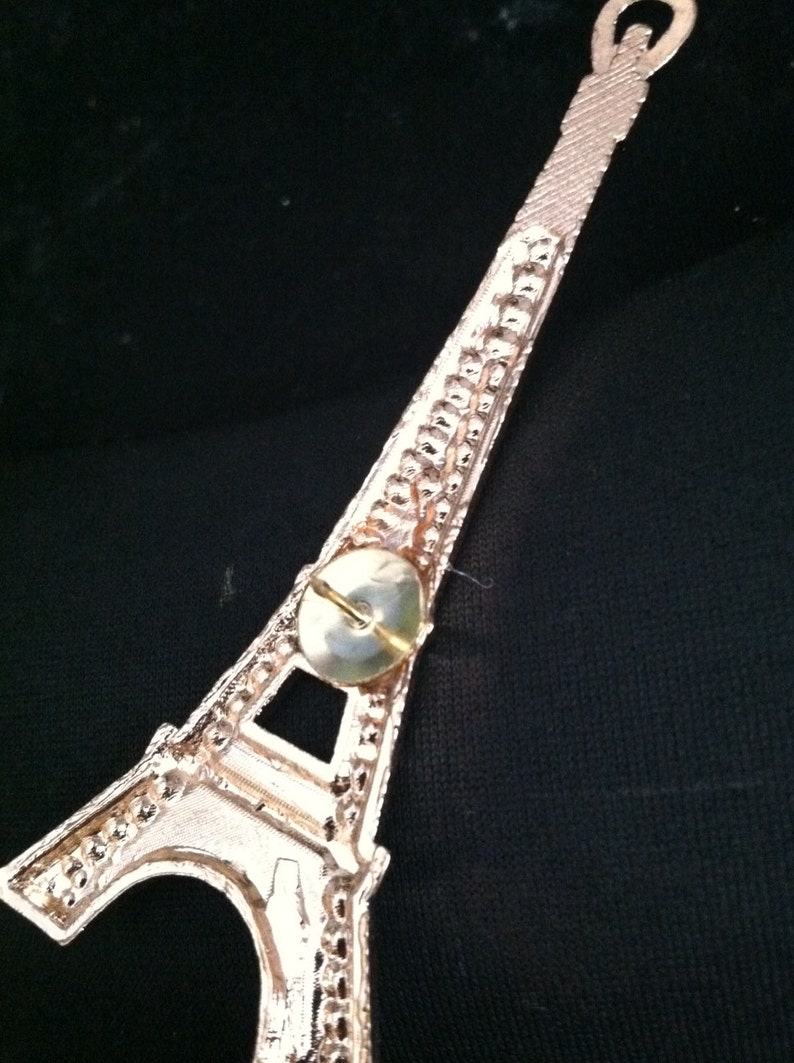 May include: A gold-colored Eiffel Tower ornament with a push pin attached to the back.