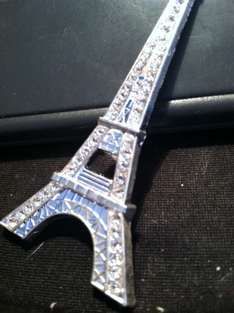 May include: A silver Eiffel Tower keychain with rhinestones. The keychain is on a black background.