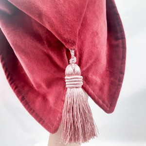 May include: A red velvet sleep mask with a pink tassel. The mask is folded in half and the tassel is hanging down.