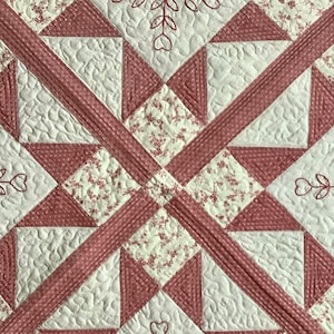 May include: A quilt with a pink and white geometric pattern. The quilt is made of squares and triangles, and it has a floral print. The quilt is quilted with a pattern of hearts and leaves.