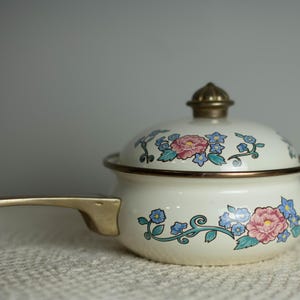 May include: A cream-colored enamel saucepan with a matching lid, featuring a floral pattern of pink roses and blue flowers with green vines. The pot has a metallic brass-toned handle and a matching brass-toned knob on the lid, with a dark rim.