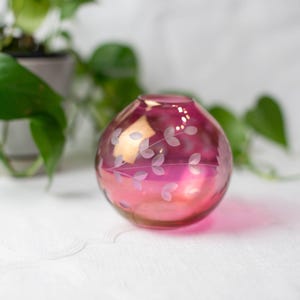 May include: A round, pink glass vase with a gradient effect, transitioning from a deep magenta at the base to a lighter pink at the top. Delicate, leaf-like patterns are etched around the vase. The vase is approximately 5 inches in diameter.