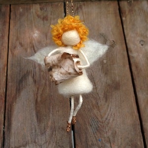 May include: A white felt angel ornament with a halo, golden hair, and a brown paper scroll in its hands. The angel is hanging from a string.