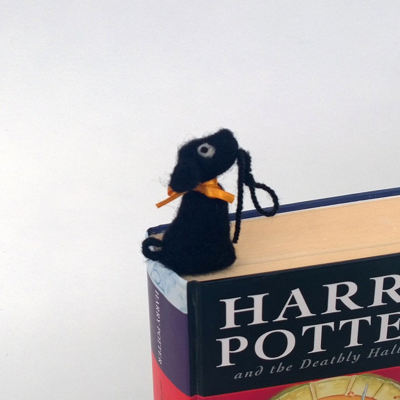 K&ouml;nnte beinhalten: Schwarzer Filz-Hunde-Lesezeichen mit gelbem Band, das auf einem Harry-Potter-Buch liegt. Der Buchtitel lautet "Harry Potter und die Heiligt&uuml;mer des Todes".