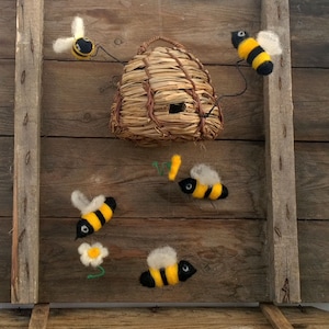 May include: A felt beehive with a straw roof and five felt bees hanging from it. The bees are yellow and black with black eyes. The beehive is hanging on a wooden wall.