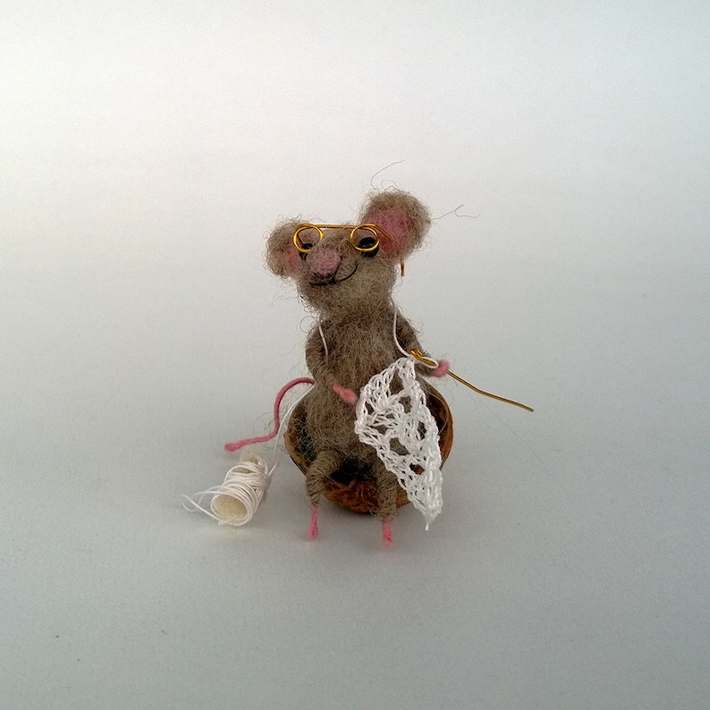 Op de afbeelding: Een kleine, grijze viltmuis met goudomrande brilletjes zit op een walnootschelp en breit een wit gehaakt doily. De muis heeft roze oren en een roze staart.