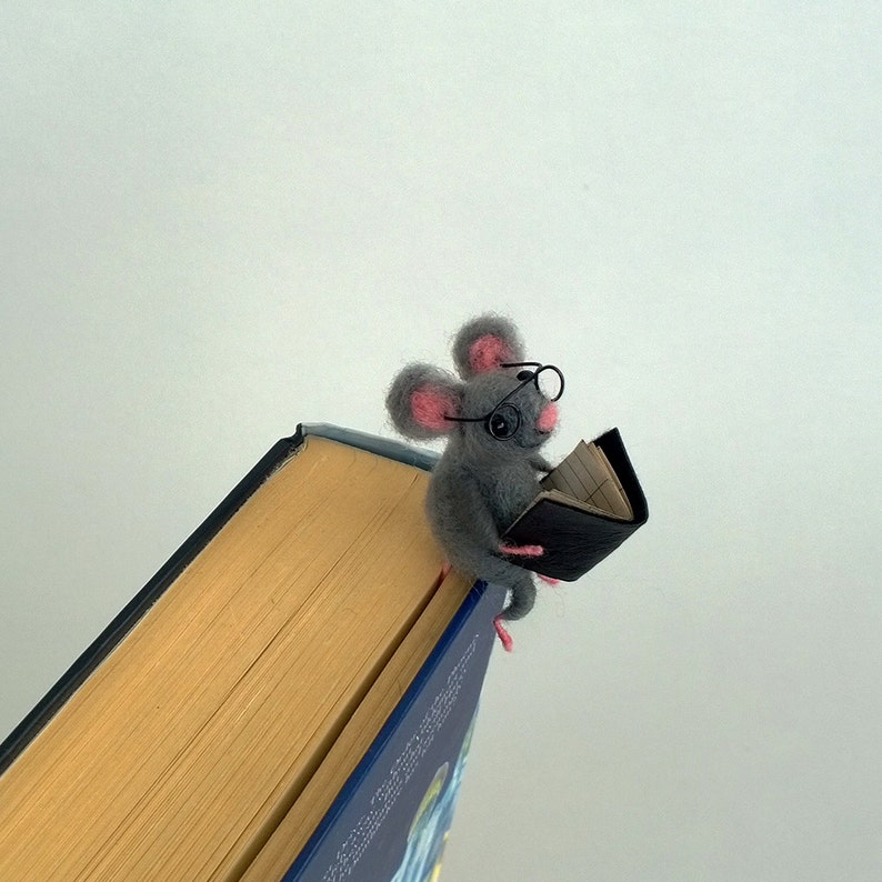 May include: A gray felt mouse wearing glasses and holding a black book bookmark. The mouse is sitting on the edge of a book.