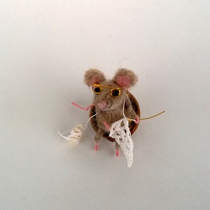 Op de afbeelding: Een kleine, bruine, naaldgevoelde muis met goudomrande bril zit in een eikel van hout en breit een wit gehaakt stuk.