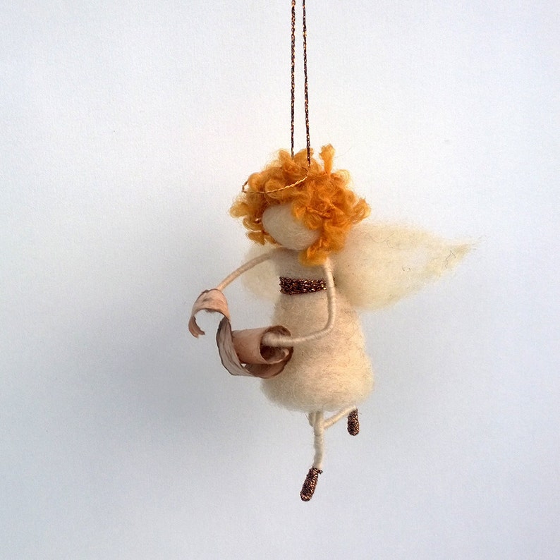 May include: A felt angel ornament with a white dress, orange hair, and a scroll in its hand. The angel is hanging from a gold string.