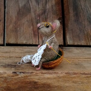 Op de afbeelding: Een kleine, grijze, vilten muis met een bril zit in een walnootschelp en breit met een wit haakgaren en een gouden naald.