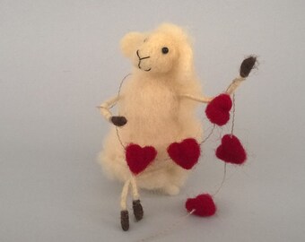 Pode incluir: Uma ovelha de feltro branco segurando uma corda de corações de feltro vermelho. A ovelha tem uma expressão doce e está sentada em um fundo branco.