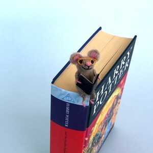 May include: A small, brown, felt mouse wearing orange glasses sits on top of a Harry Potter book. The mouse is holding a small, black book. The book cover reads "Harry Potter and the Half-Blood Prince."