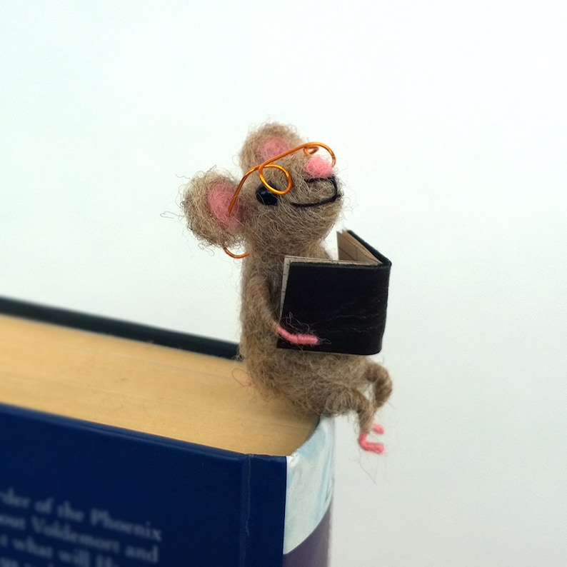 May include: A brown felt mouse wearing orange glasses and holding a black book. The mouse is sitting on the top of a book.