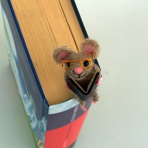 May include: A brown and gray felt mouse wearing orange glasses and holding a small black book. The mouse is perched on the edge of a book with a blue and red cover.