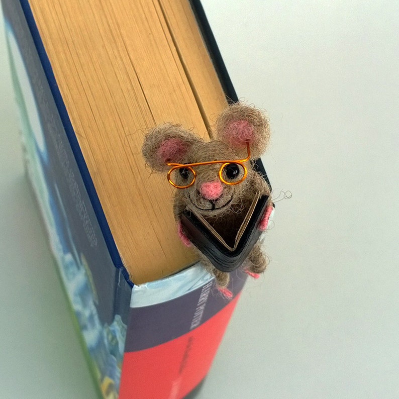 May include: A brown and gray felt mouse wearing orange glasses and holding a small black book. The mouse is perched on the edge of a book with a blue and red cover.