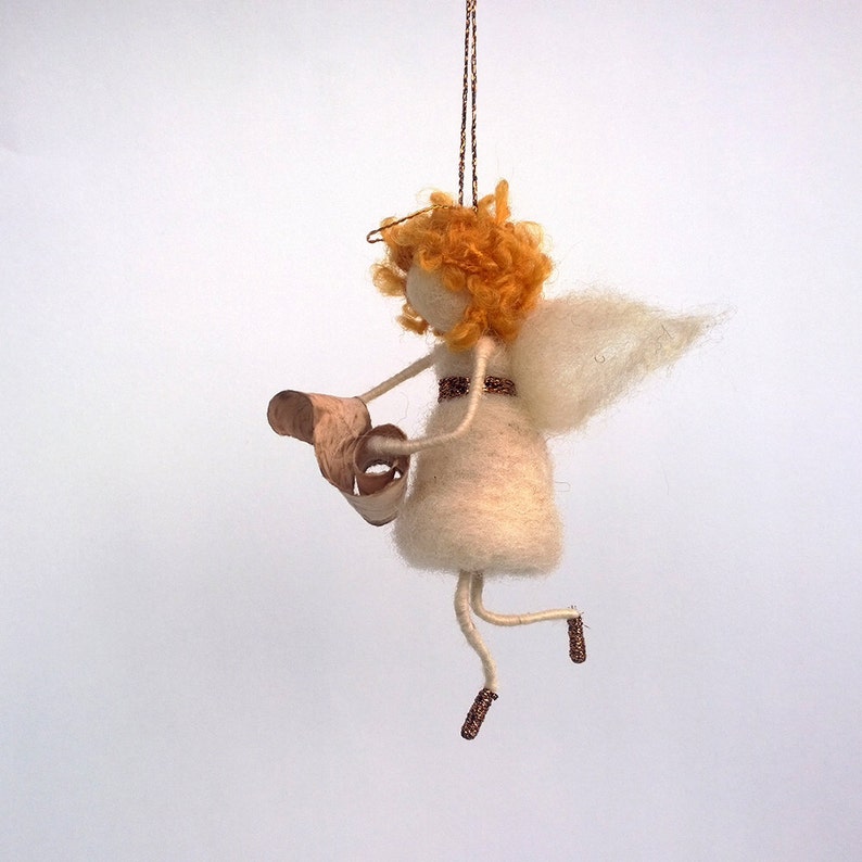 May include: A white felt angel ornament with a gold wire halo and orange curly hair. The angel is holding a brown scroll in its hands and has a gold ribbon around its waist. The angel is hanging from a gold string.