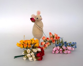 Puede incluir: Un ratón de fieltro blanco sosteniendo una sola rosa roja, rodeado de cuatro ramos de pequeñas rosas de papel en amarillo, naranja, azul y rosa.