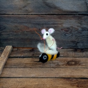Peut inclure: Une sculpture en feutre fantaisiste représentant une souris blanche chevauchant un bourdon. L'abeille a une tête noire, un corps rayé jaune et noir et des pattes en fil de fer. La souris a des oreilles roses et une longue queue. Fond en bois rustique.