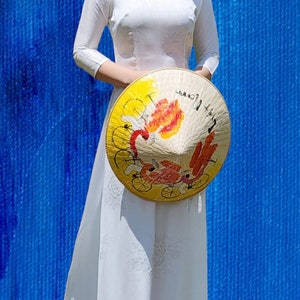 May include: A woman wearing a white dress and sunglasses holds a traditional Vietnamese conical hat with a colourful design. The hat is decorated with a red, yellow and orange floral pattern.