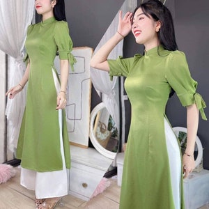 Può includere: Un vestito verde con un pannello laterale bianco e maniche corte. Il vestito ha un colletto alla coreana e una silhouette aderente. La modella indossa pantaloni bianchi.