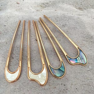 May include: Five gold-toned hair pins with decorative ends. Two pins have white, pearlescent inlays, and two have iridescent, abalone-like inlays. The pins are arranged on a textured, light gray surface.