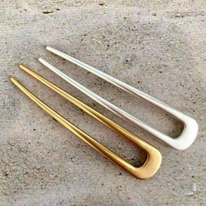 May include: Two hair pins, one gold-toned and one silver-toned, are displayed on a textured gray surface. The pins have a U-shaped design with three prongs. The gold pin is on the left, and the silver pin is on the right.