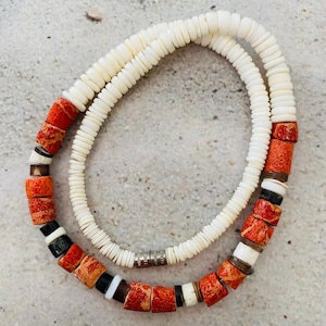 May include: A necklace featuring a long strand of small, flat, white disc beads and a shorter section of larger, textured, orange-red cylindrical beads interspersed with smaller black, white, and metallic brown spacer beads and a metal clasp.