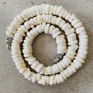 May include: A necklace composed of numerous off-white, disc-shaped beads with a porous texture. The beads are strung together in a circular arrangement, featuring small metallic silver-toned cylindrical clasps at both ends of the strand.