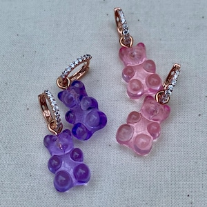 May include: A pair of earrings with gummy bear charms. One earring has a purple gummy bear charm and the other has a pink gummy bear charm.