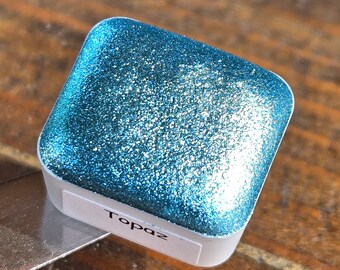 May include: A small square container of blue glitter paint with the word "Topaz" written on the label.