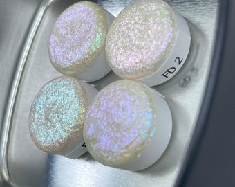 May include: Four iridescent white round glass cabochons with a glittery finish. The cabochons are arranged in a square shape. The cabochon in the top right corner has the text "FD 2" written on it.