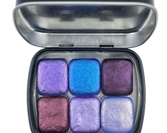 May include: A metal tin with six pans of watercolor paint. The pans are arranged in two rows of three. The colors are purple, blue, dark purple, burgundy, lavender, and light blue.