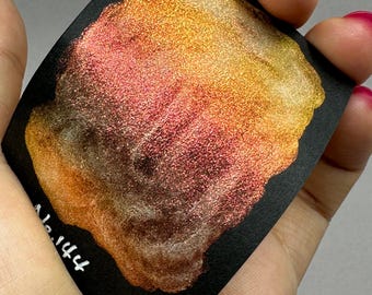 May include: A close-up of a metallic gold, orange, and red shimmery paint swatch on a black surface. The swatch has a textured, glittery finish.