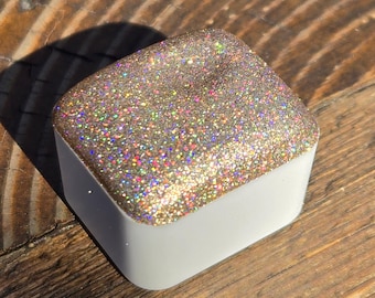 May include: A small square container filled with iridescent gold glitter. The container is white with a smooth, glossy finish.