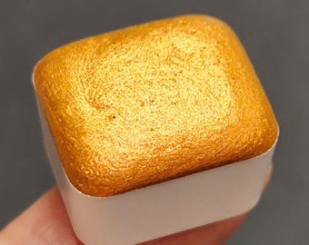 May include: A gold watercolor paint pan in a white plastic container. The paint has a metallic sheen.