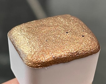 May include: A close-up of a gold watercolor pan with a metallic sheen. The pan is in a white plastic container.