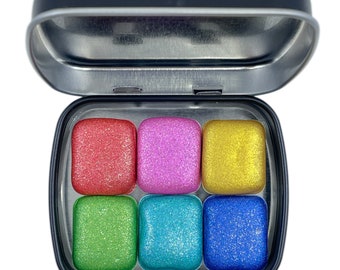 May include: A tin with six small, square, shimmery eyeshadows. The colors are red, pink, yellow, green, blue, and dark blue.