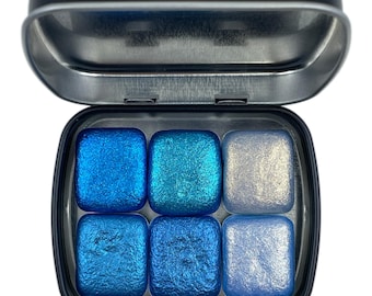 May include: An open, black metal tin filled with six square watercolour paint pans. The colours are various shades of blue and silver, with a shimmering, metallic finish. The paints range from deep blue to light silver.