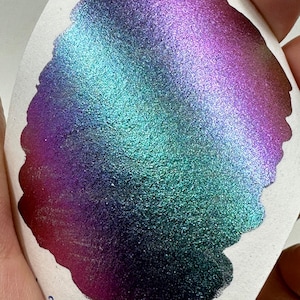 May include: A close-up of a shimmery, iridescent teal and purple paint sample. The paint has a metallic sheen and a textured finish.
