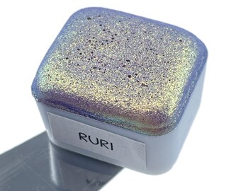 May include: A small square container of iridescent blue and gold glitter with the word "RURI" printed on the label.