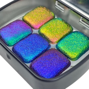 May include: Six iridescent glass tiles in a metal tin. The tiles are in shades of green, blue, and purple.